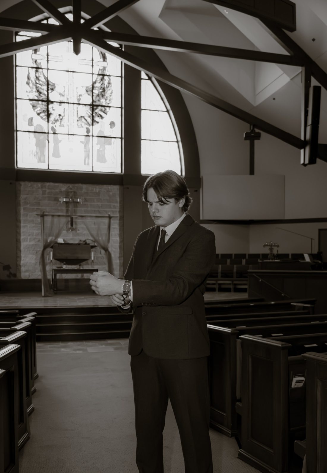 Zack Riordan in a dark suit, fastening his cuff in a church sanctuary beneath a stained-glass window